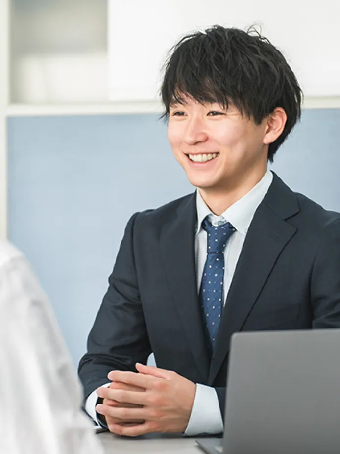 デスクで笑顔で打ち合わせをするスーツ姿の男性社員の写真